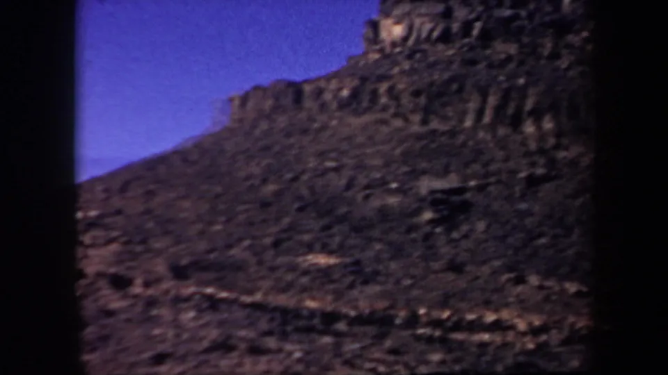 1957 Arizona 8mm frame showing a steep rocky slope with cliff-like outcrops and blue-purple sky.