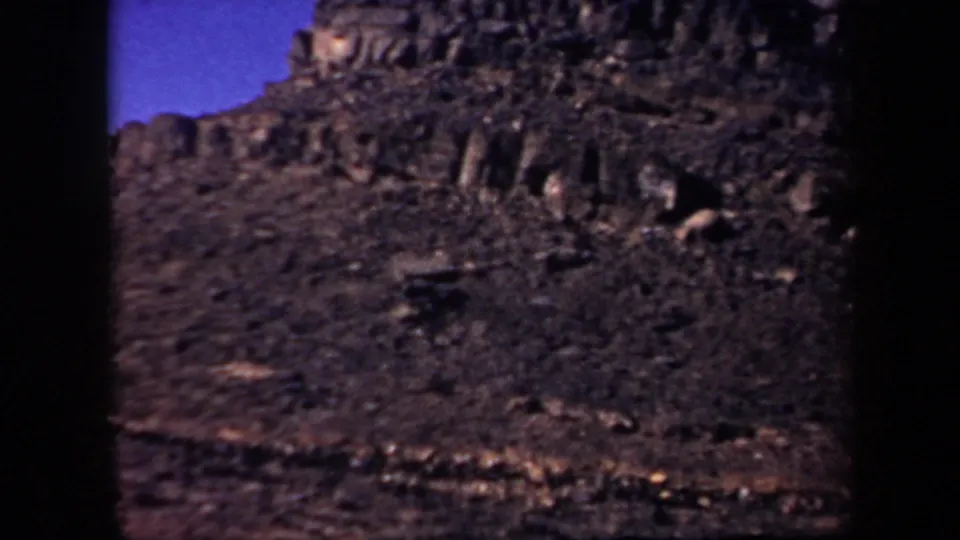 1957 Arizona 8mm frame showing a rugged brown mountainside with dark jagged rock formations and a narrow band of blue sky.