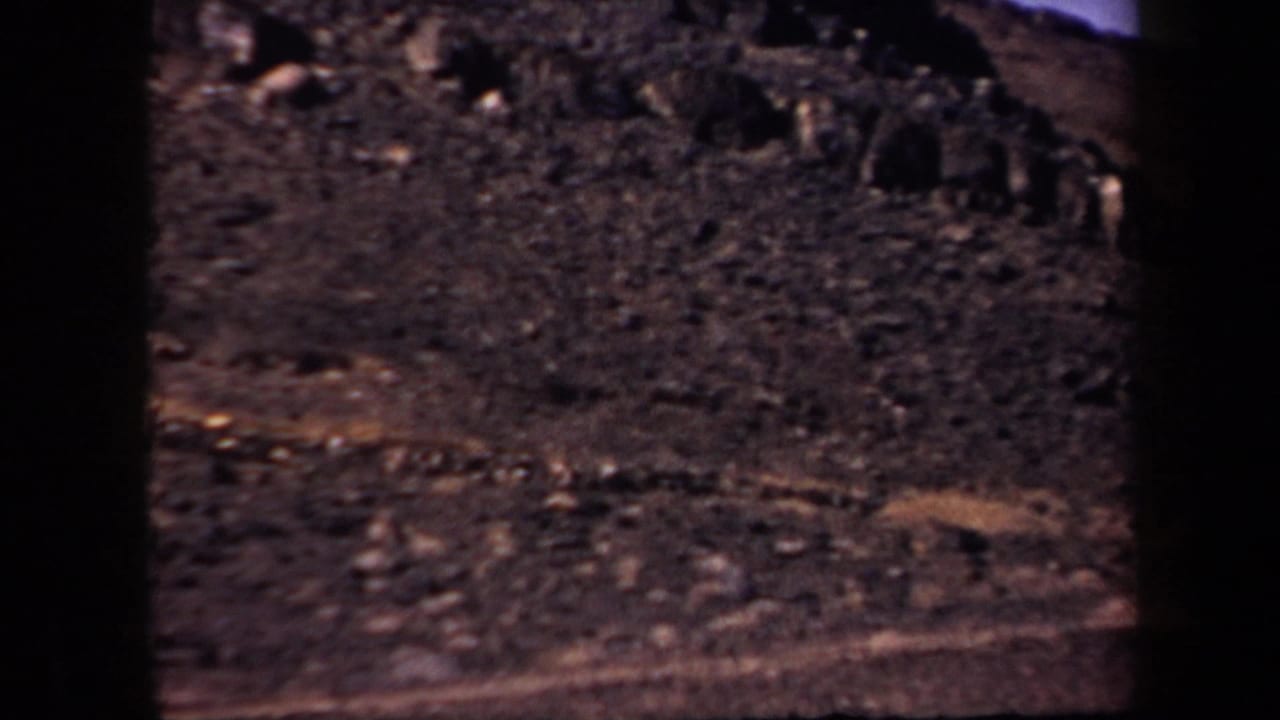 1957 Arizona home-movie frame with a steep arid slope, dark rock clusters, scattered boulders, and a faint path across the mountainside.