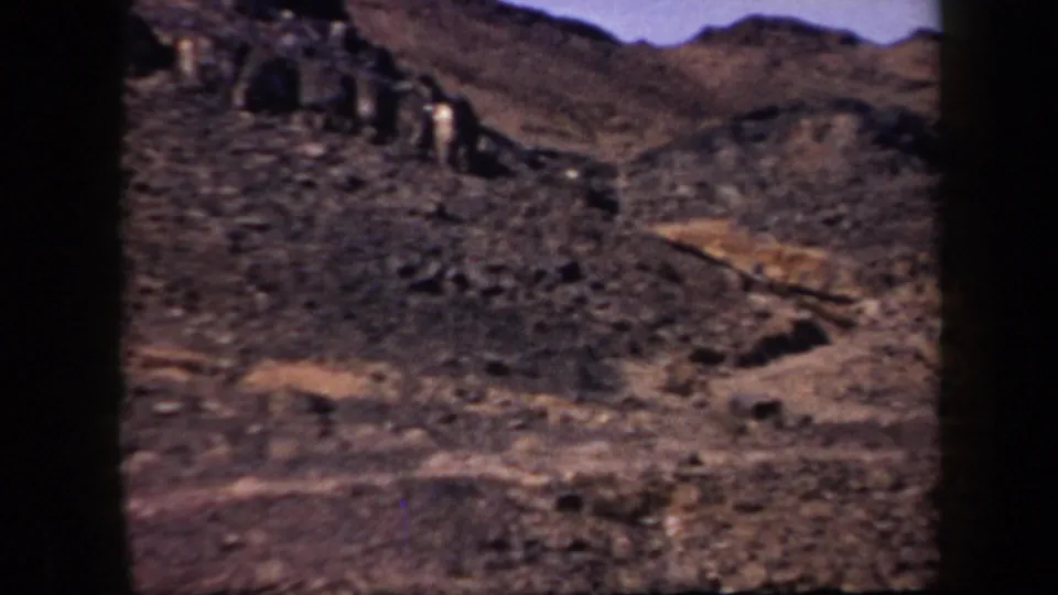 1957 Arizona 8mm frame showing a dry mountainside with layered rocky ridges, loose stones, and pale tracks crossing the slope.