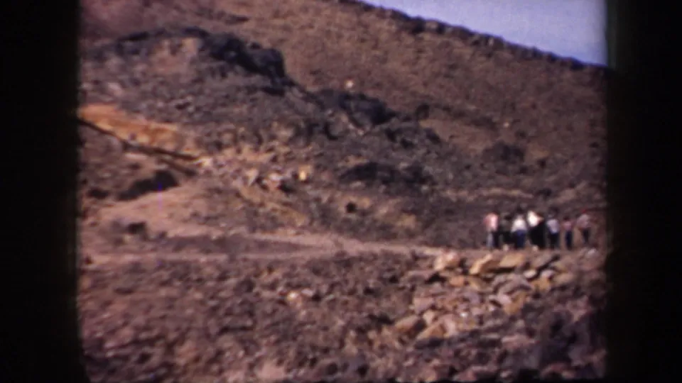 1957 Arizona home-movie frame showing a rocky mountainside and a small distant group of people standing near a pale path.