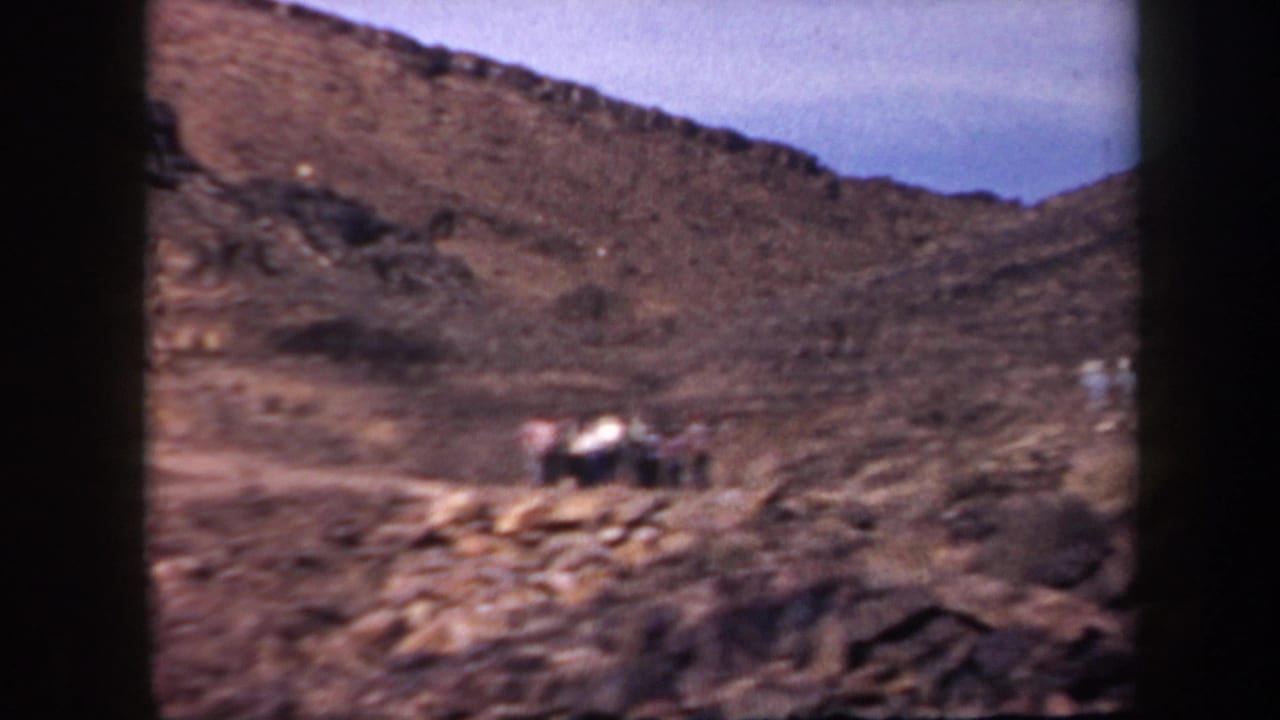 Rocky desert hillside with small adults gathered on the slope under a pale sky.