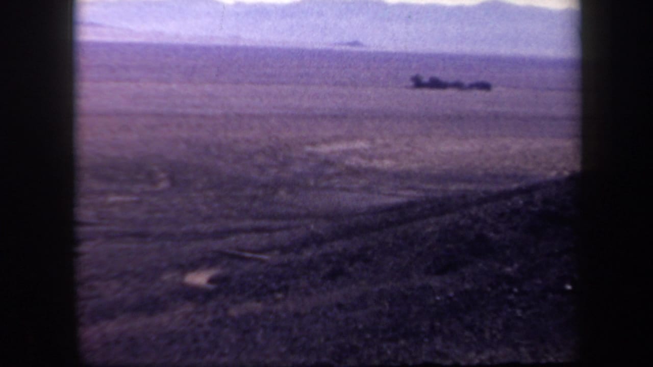 Wide desert plain or slope with distant dark shapes near the horizon.