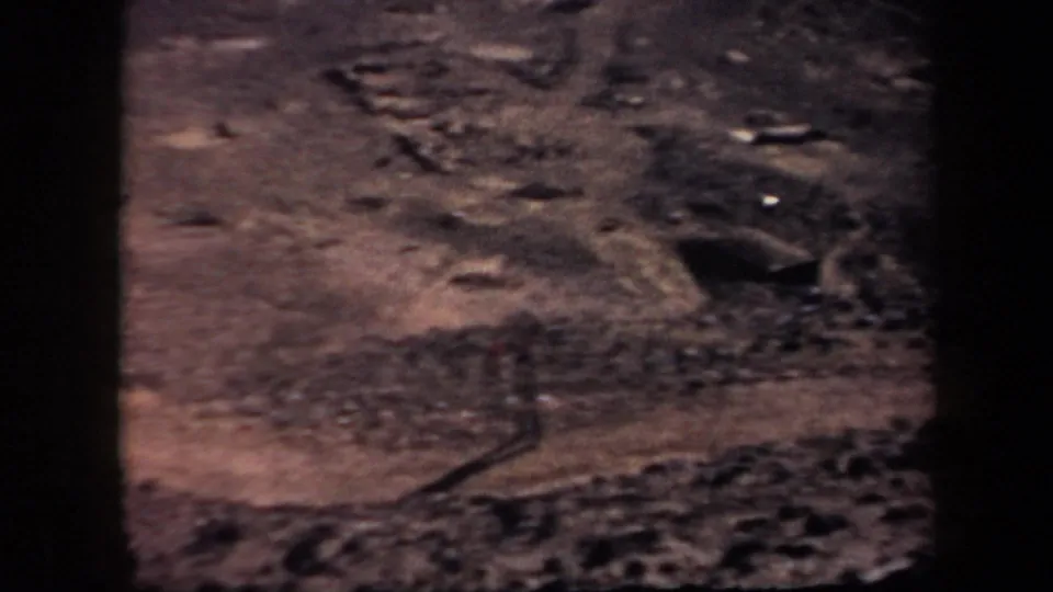 Color-faded 8mm frame of rough Arizona desert terrain with stones, tracks, and shallow depressions.