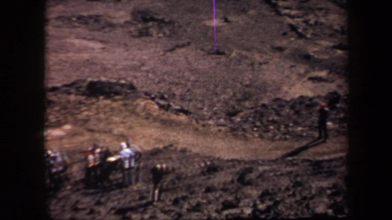 In 1957 Arizona home-movie footage, several people stand on dusty uneven terrain near a rough dirt racetrack area.