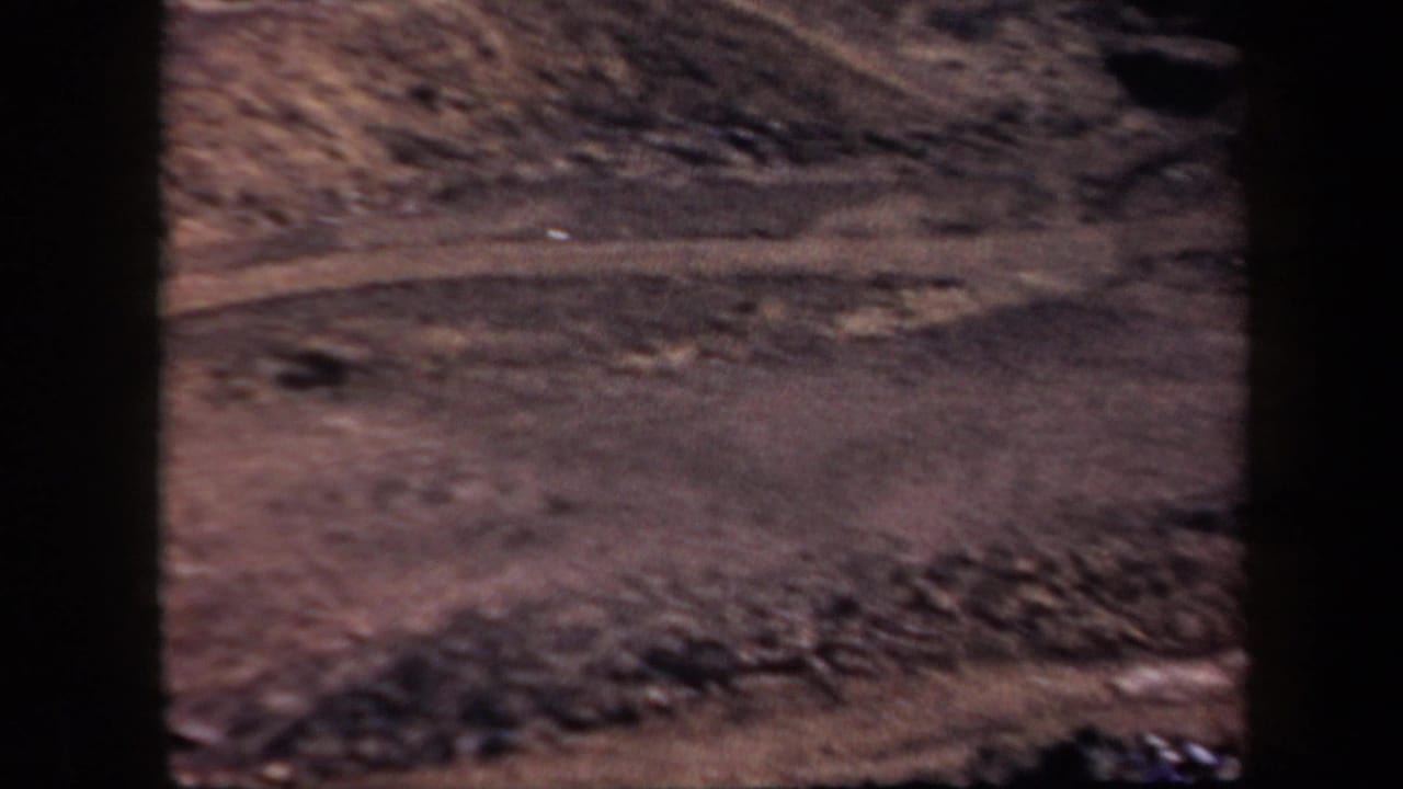 A dusty curved dirt road cuts across a barren hillside with no people clearly visible.