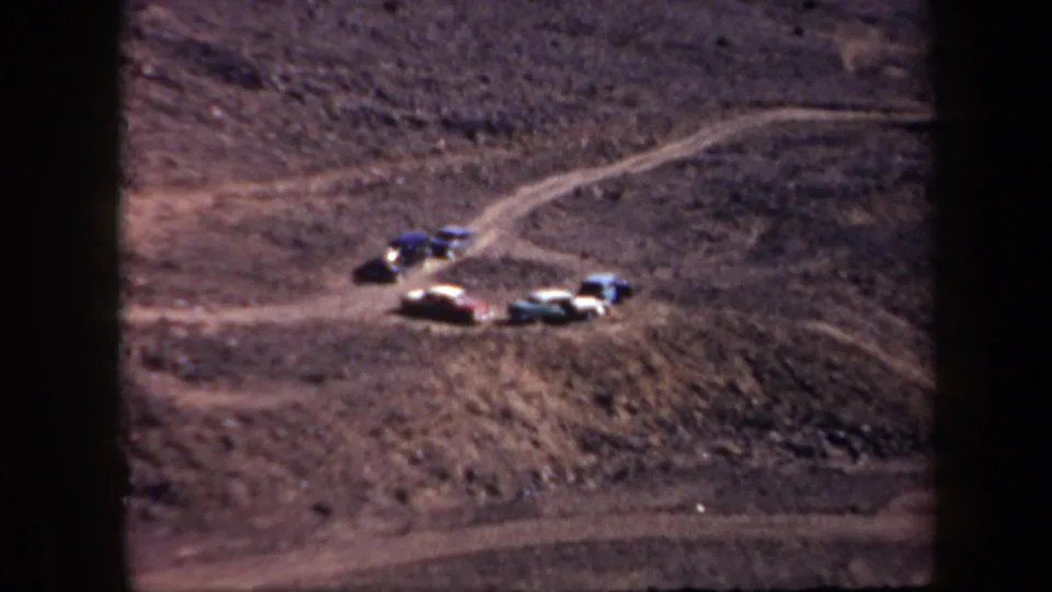 Several 1950s-era cars sit near a winding dirt road in Arizona, with open hills surrounding the racetrack area.