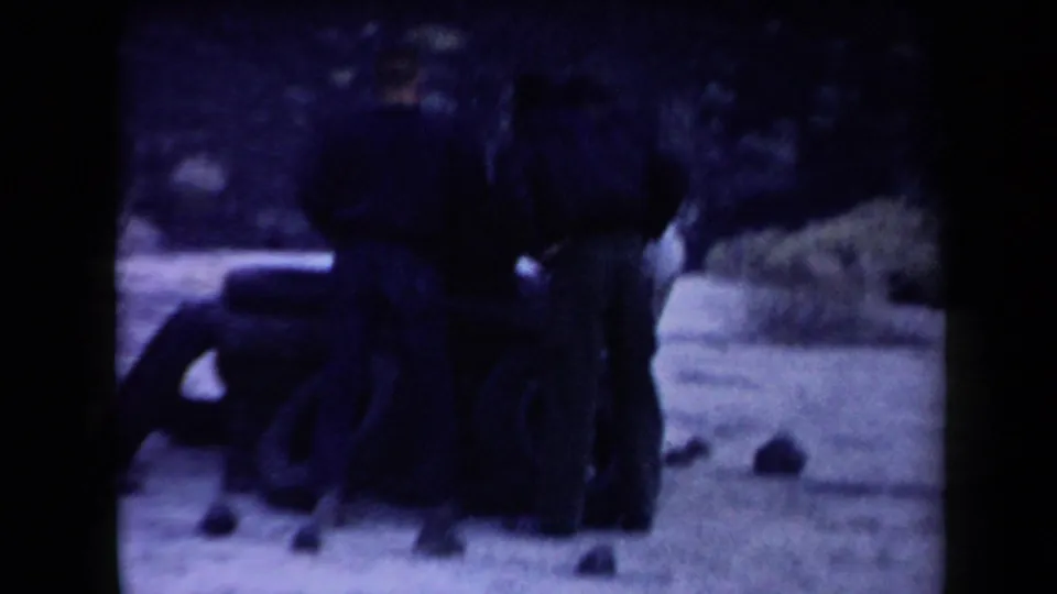 Three men in 1958 Arizona home-movie footage gather beside a low tire stack in a dry, shrub-dotted landscape.