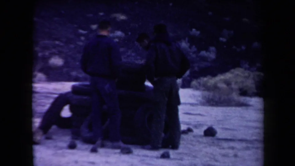 In the Arizona desert scene, three men stand close to a small tire tower with sparse brush and rocks behind them.