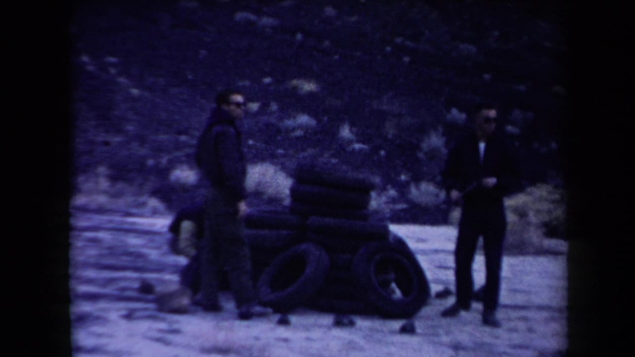 Three men in casual 1950s clothing stand near a pile of rubber tires in an Arizona desert setting.