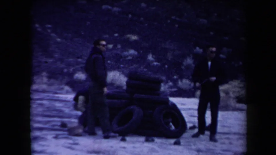 Three men in casual 1950s clothing stand near a pile of rubber tires in an Arizona desert setting.