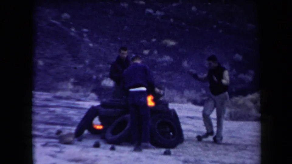 A small orange flame appears at the tire pile while men stand, crouch, and gesture in the desert scene.