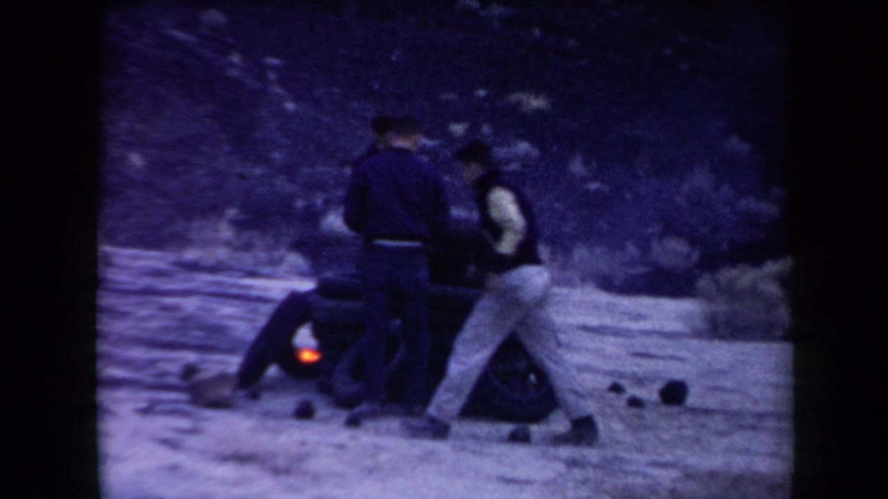 In supplied 1958 Arizona 8mm footage, three adult men gather around stacked tires as a small fire burns in a sparse desert setting.