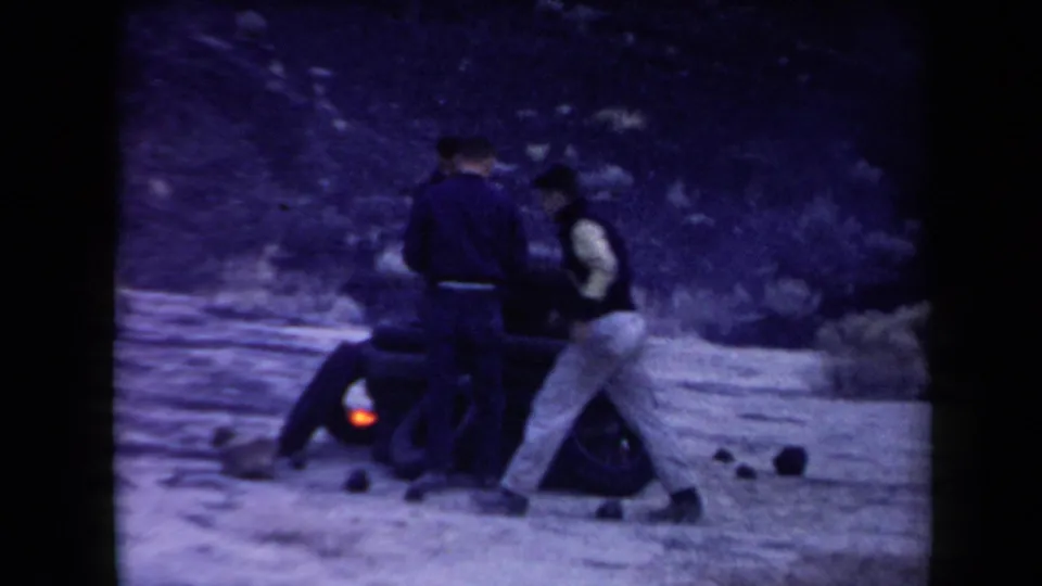 In supplied 1958 Arizona 8mm footage, three adult men gather around stacked tires as a small fire burns in a sparse desert setting.