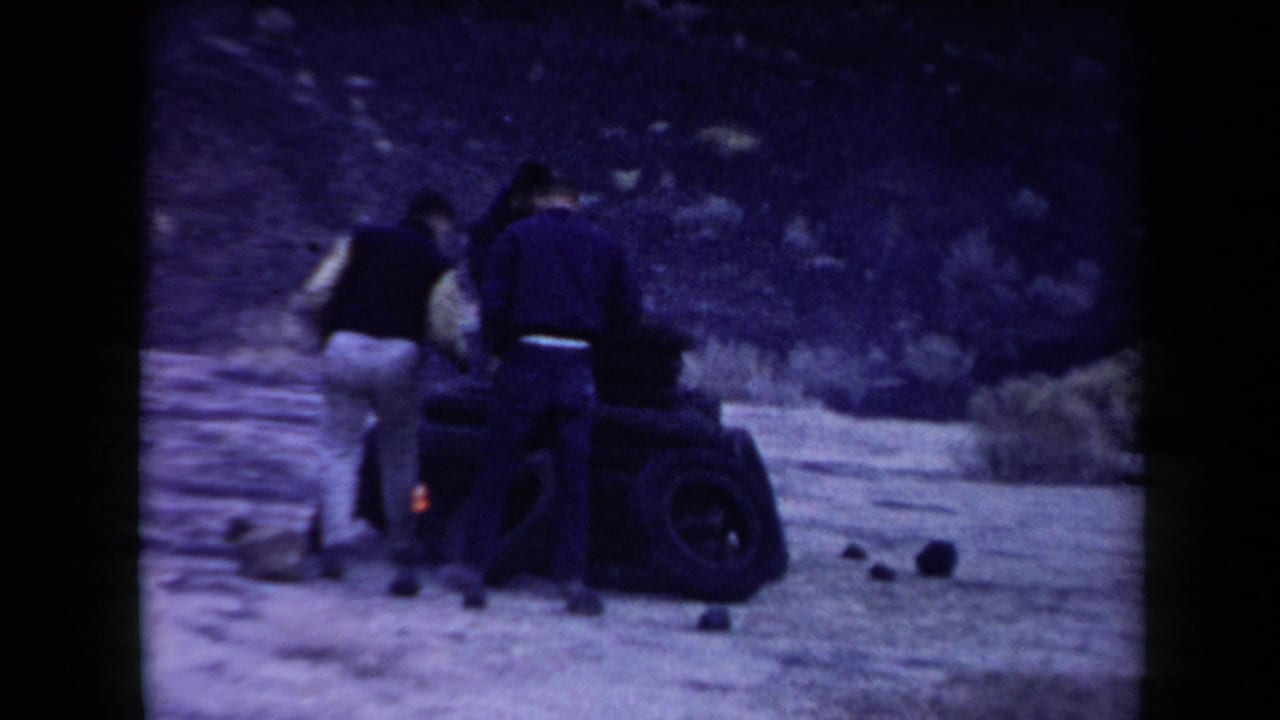 Three men in mid-century clothing appear around a burning tire pile in the Arizona desert, with scrub and a rocky slope behind them.