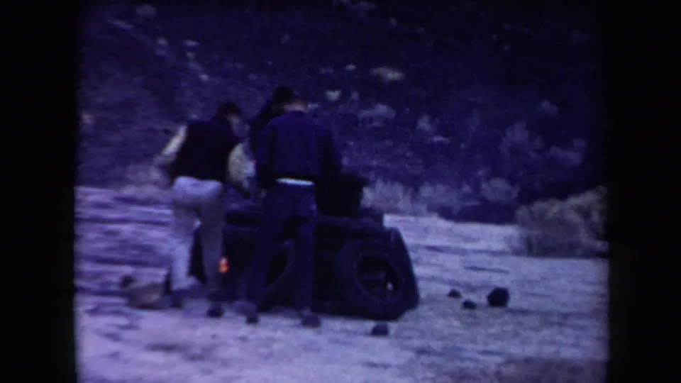 Three men in mid-century clothing appear around a burning tire pile in the Arizona desert, with scrub and a rocky slope behind them.