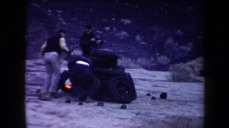 In the purple-blue 8mm frame, men work close to a burning tire pile on pale desert ground with scattered rocks nearby.
