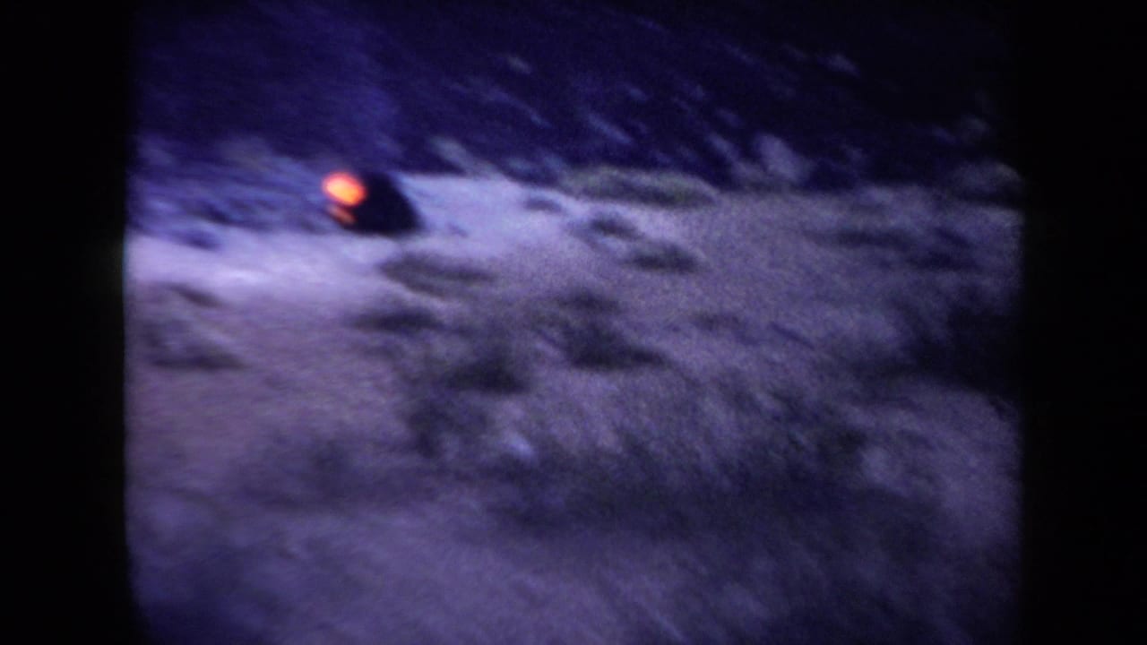 1958 Arizona 8mm frame showing dark mountainous ground with a small orange glow and strong purple-blue film color shift.