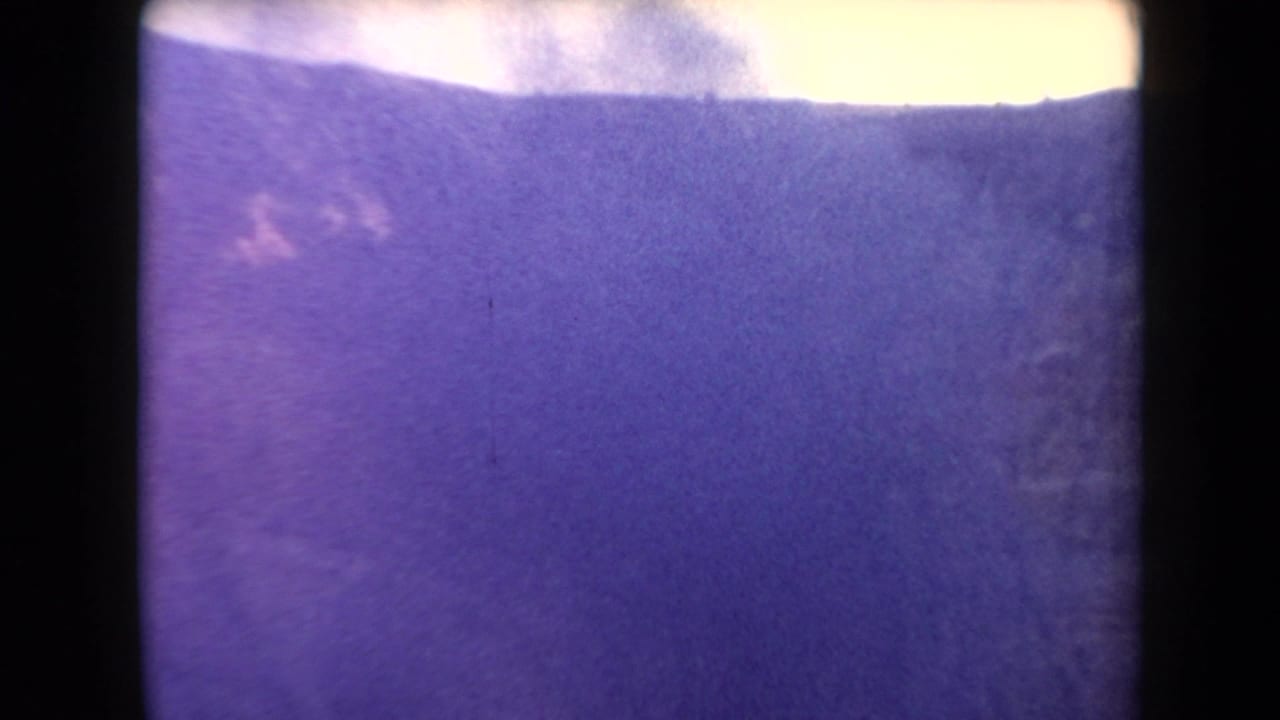 1958 Arizona home-movie frame with a dark ridge line, pale sky, and light smoke drifting above winter-toned terrain.