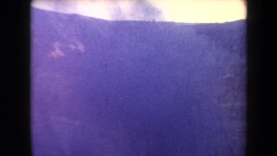 1958 Arizona home-movie frame with a dark ridge line, pale sky, and light smoke drifting above winter-toned terrain.