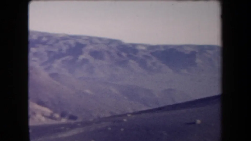 1958 Arizona 8mm frame showing a distant arid canyon landscape with eroded ridges, talus slopes, pale sky, and a dark foreground line.