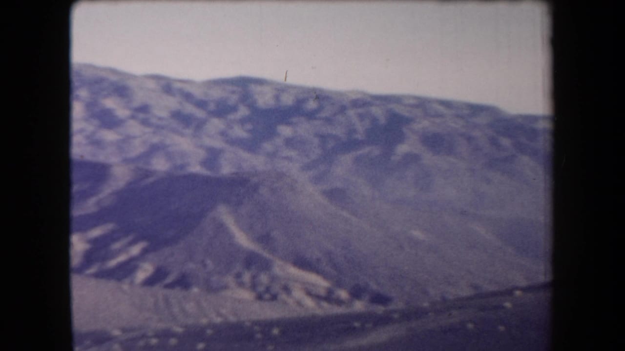1958 Arizona home-movie frame showing arid mountain ridges and sparse desert ground under a washed pale sky.