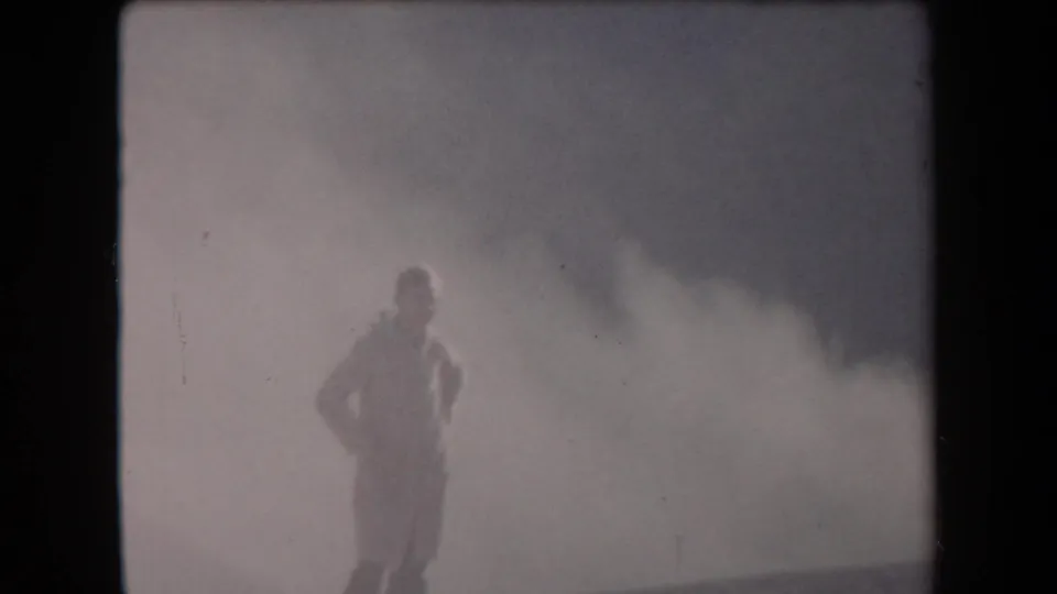 A 1958 Arizona 8mm frame shows a lone uniformed figure against a large smoke plume and blurred terrain.