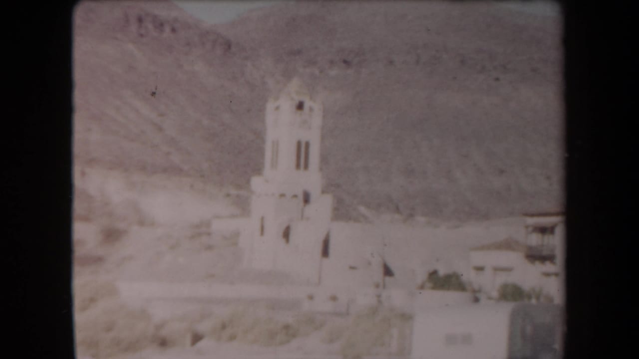 White stucco bell tower against desert mountains with buildings at the right edge.