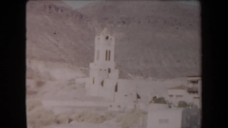 1958 Arizona home-movie frame showing a white stucco bell tower and adjacent desert-town buildings below arid mountains.