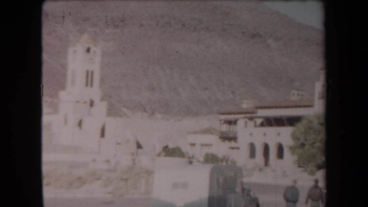 1958 Arizona frame with a bell tower, arched stucco building, desert hillside, and two distant adults in the foreground area.