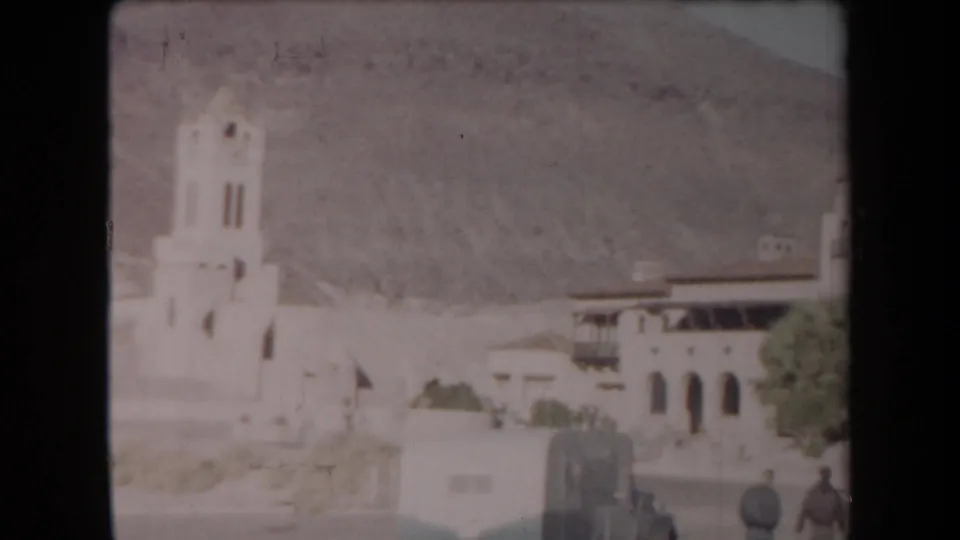 1958 Arizona frame with a bell tower, arched stucco building, desert hillside, and two distant adults in the foreground area.