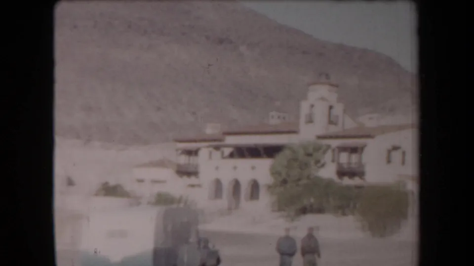 Arizona 1958 home-movie view of a stucco town building with arched openings, a parked car, shrubs, and two adults walking in front.