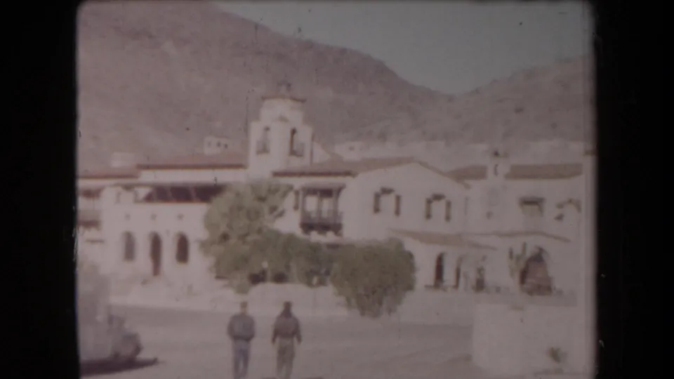 Wide 1958 Arizona exterior showing stucco buildings with a bell tower, two adults in the street area, a car, trees, and desert hills.