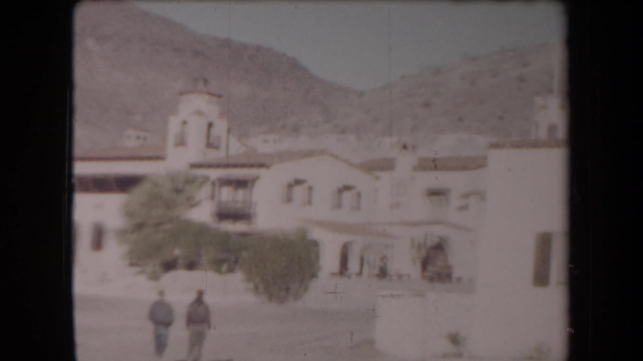 Pale adobe-style building below desert mountains with two men walking in the courtyard.