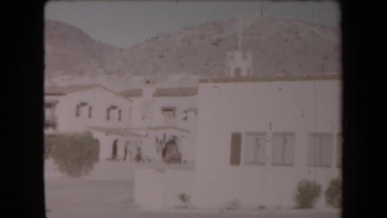 Arizona desert home-movie view of a pale stucco structure with arched details, courtyard, trees, and low mountains behind it.