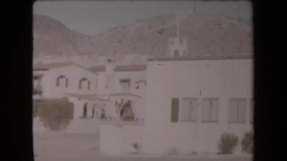 Arizona desert home-movie view of a pale stucco structure with arched details, courtyard, trees, and low mountains behind it.
