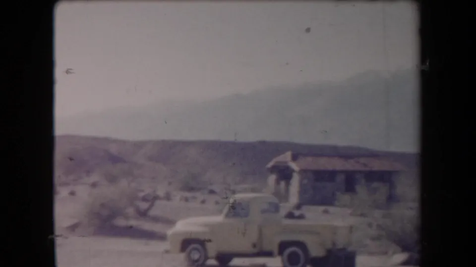 1958 Arizona frame showing a yellow pickup truck near a small rustic building, scrub vegetation, and distant desert hills.