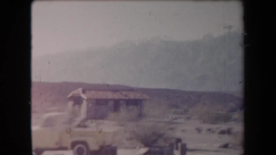 Soft 8mm frame of a yellow pickup and small desert building in Arizona, with scrub, open ground, and mountain slopes beyond.
