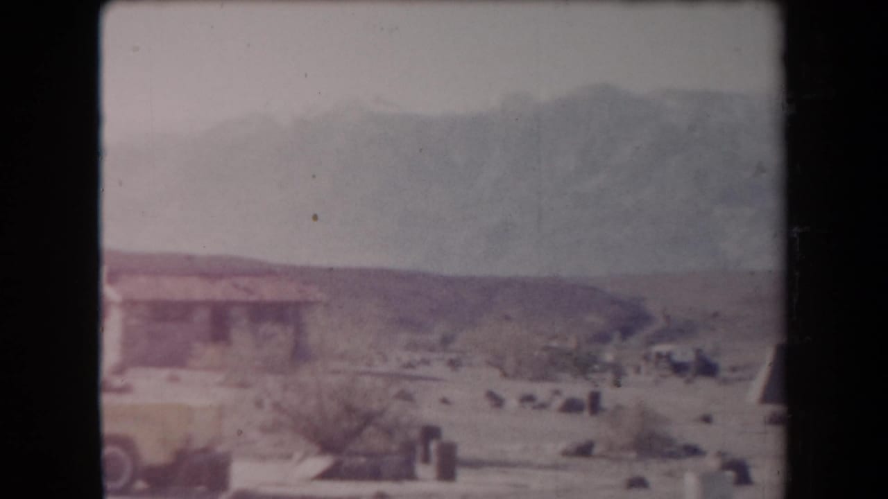1958 Arizona home-movie frame showing a small desert structure, yellow vehicle, dry brush, pale sky, and distant mountains.