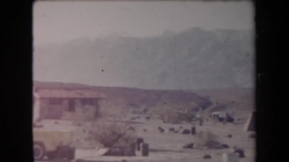 1958 Arizona home-movie frame showing a small desert structure, yellow vehicle, dry brush, pale sky, and distant mountains.