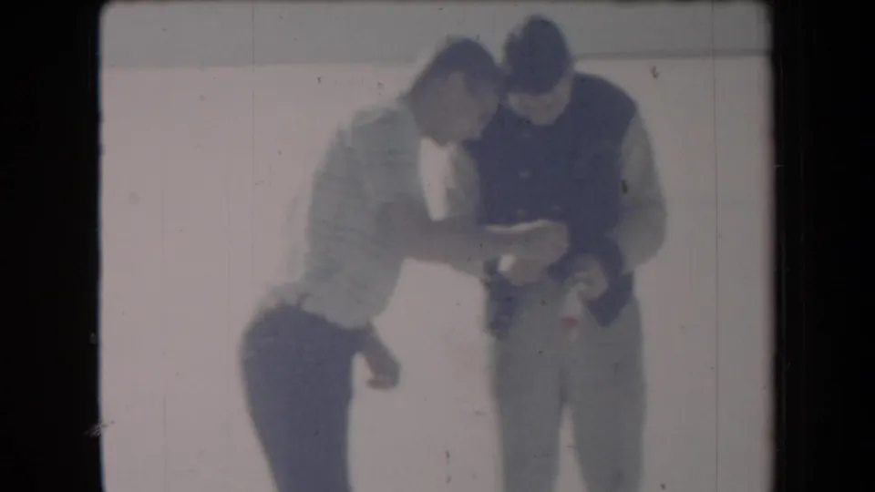 In faded 8mm color, two men stand in bright outdoor light and examine a small object during a home-movie moment.