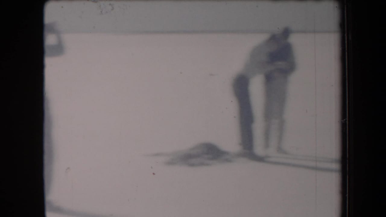 A teen bends near a football on the bright flat ground as another figure stands nearby.