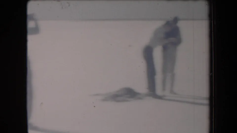 Home-movie frame from Arizona in 1958 showing a teen bending toward a football on a bright salt-flat surface, with another person at the edge of the scene.