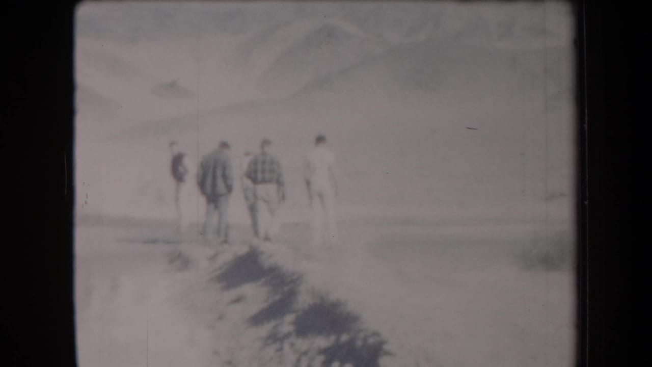 Several men walk away along a pale desert path with mountains in the background.