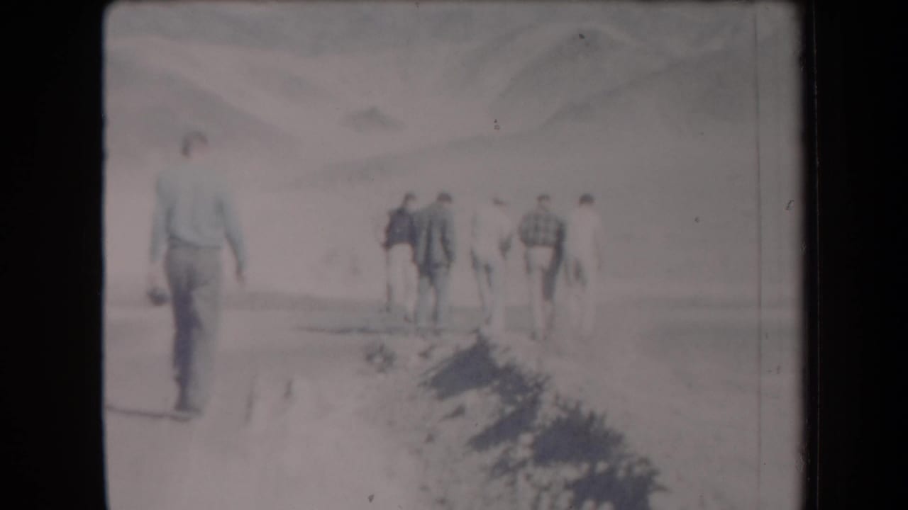 1958 Arizona home-movie frame with one adult man closer to camera and several men ahead on a desert track near mountains.