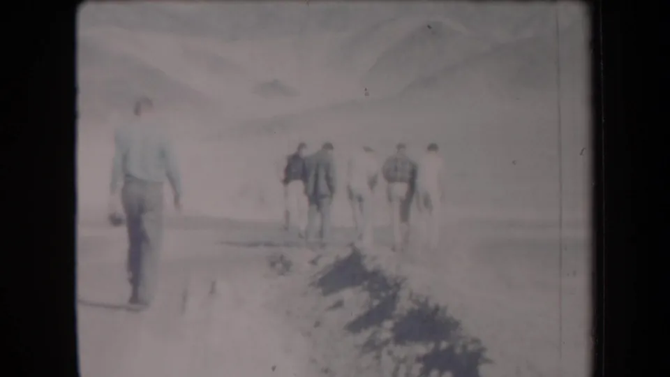 1958 Arizona home-movie frame with one adult man closer to camera and several men ahead on a desert track near mountains.