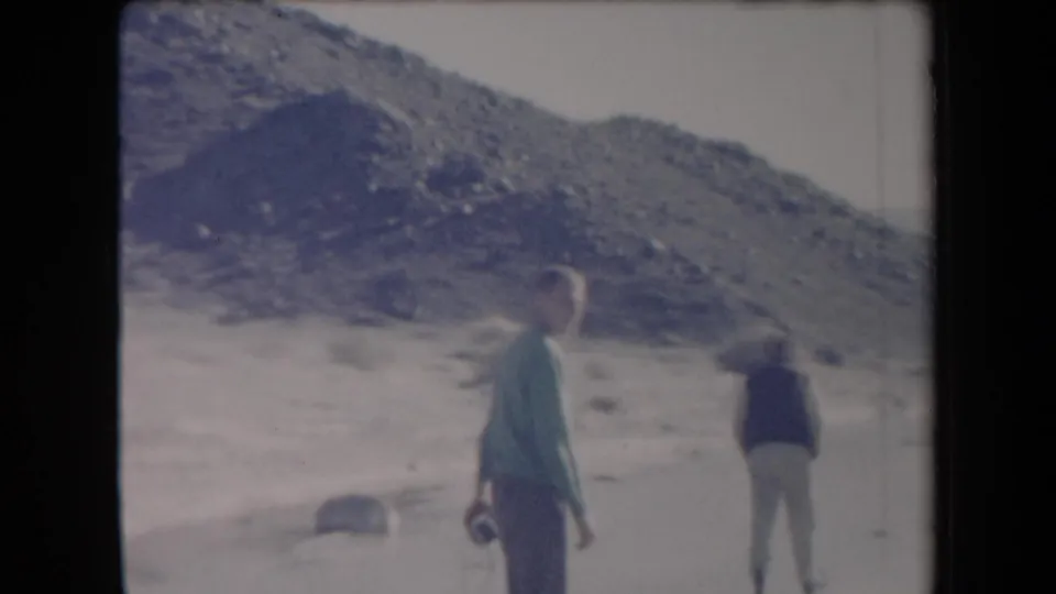 1958 Arizona 8mm frame showing two adult men crossing sandy desert terrain near a rocky hill.