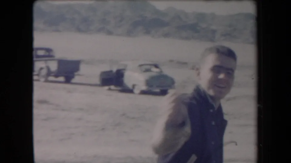 Adult man near a pickup, trailer, and car in a 1958 Arizona desert home-movie frame.