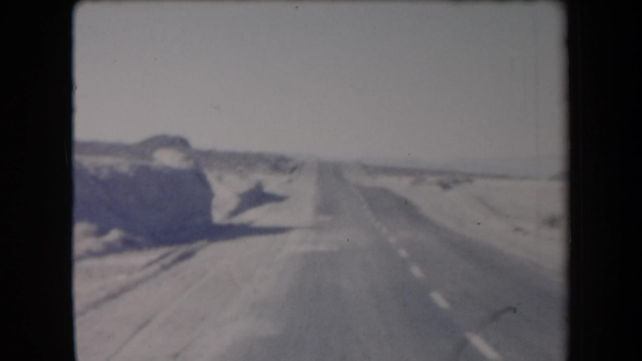 1958 Arizona road-trip frame with a straight paved highway, dry shoulders, roadside hills, and a hazy desert horizon.