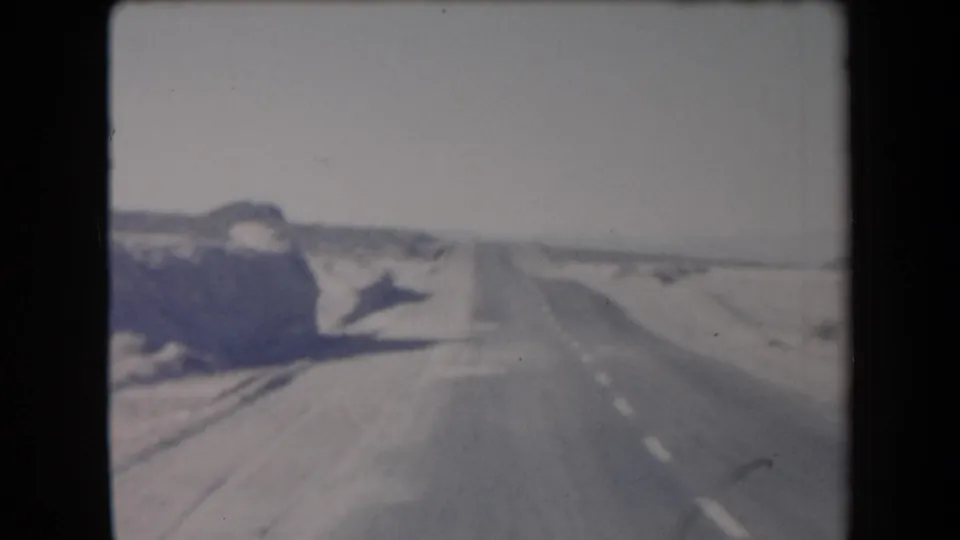 1958 Arizona road-trip frame with a straight paved highway, dry shoulders, roadside hills, and a hazy desert horizon.